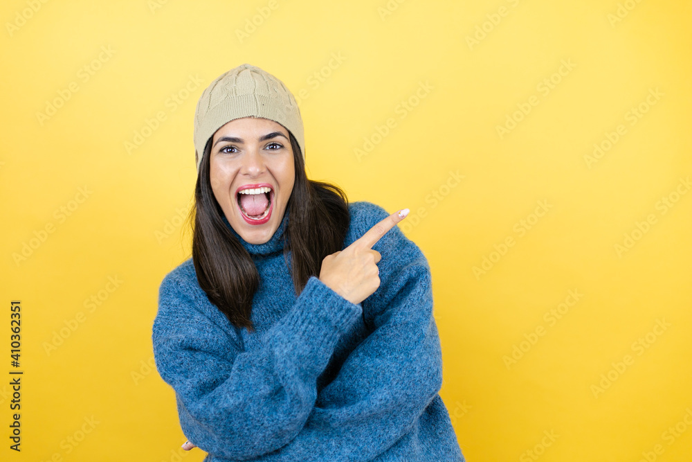 Young beautiful woman wearing blue casual sweater and wool hat smiling and pointing with hand and finger to the side