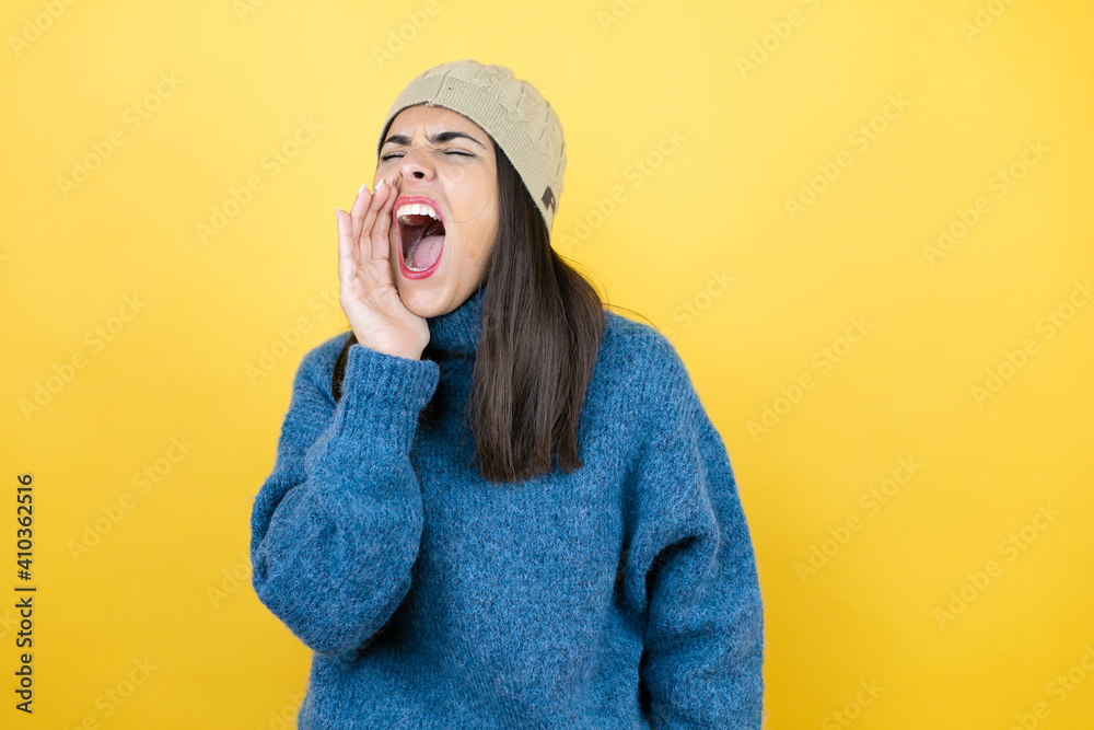 Young beautiful woman wearing blue casual sweater and wool hat shouting and screaming loud to side with hand on mouth Young beautiful woman wearing blue casual sweater and wool hat shouting and screaming loud to side with hand on mouth