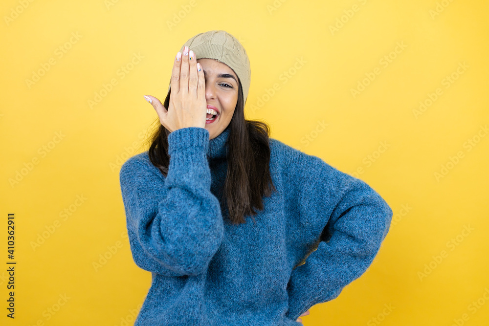 Young beautiful woman wearing blue casual sweater and wool hat covering one eye with hand, confident smile on face and surprise emotion. Young beautiful woman wearing blue casual sweater and wool hat covering one eye with hand, confident smile on face and surprise emotion.