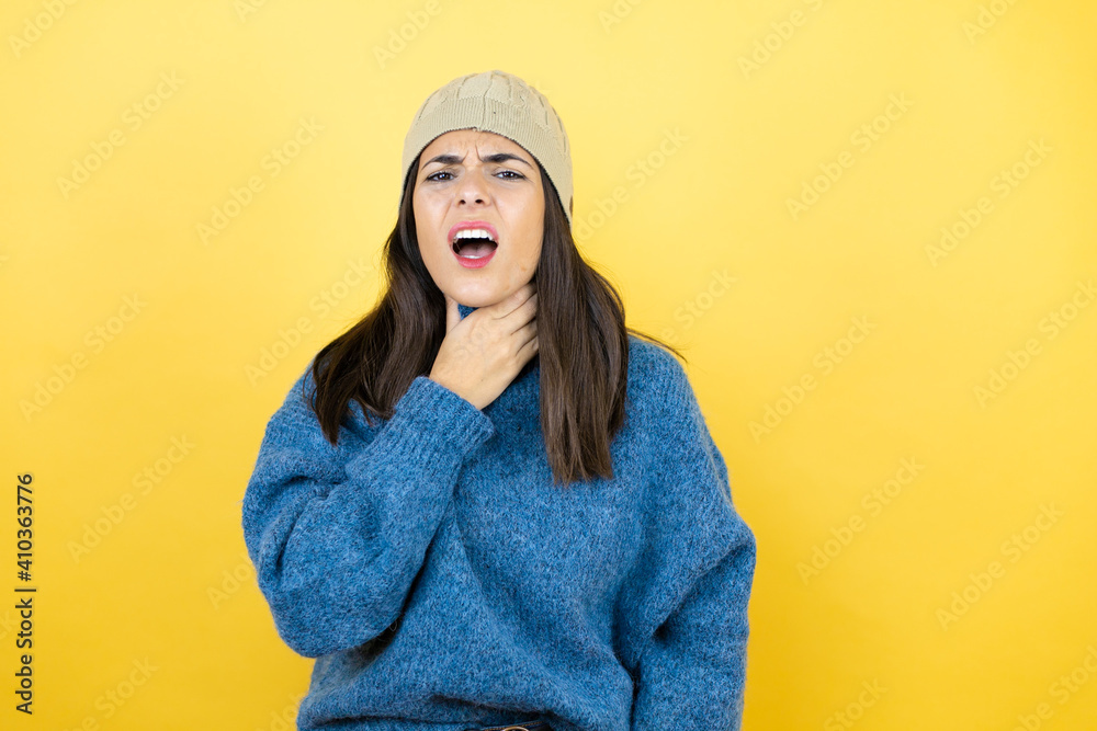 Young beautiful woman wearing blue casual sweater and wool hat touching painful neck, sore throat for flu, clod and infection Young beautiful woman wearing blue casual sweater and wool hat touching painful neck, sore throat for flu, clod and infection