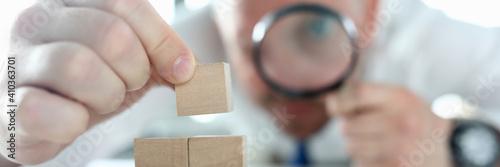 Close-up of man looking on wooden cubes stack through magnifying glass. Concentrated person building pyramid on working table. Business strategy and smart decision concept photo