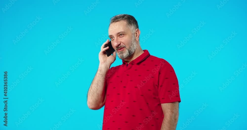 Portrait of happy Caucasian middle-aged male in red t-shirt speaking on smartphone in good mood while standing on blue background alone in studio, talking on cellphone, conversation concept Portrait of happy Caucasian middle-aged male in red t-shirt speaking on smartphone in good mood while standing on blue background alone in studio, talking on cellphone, conversation concept