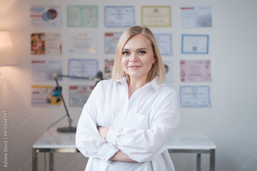 Portrait of blonde woman doctor in a beauty salon in white uniform, beautician looking at the camera and smiling, master of permanent makeup stands on a background of certificates, selective focus Portrait of blonde woman doctor in a beauty salon in white uniform, beautician looking at the camera and smiling, master of permanent makeup stands on a background of certificates, selective focus