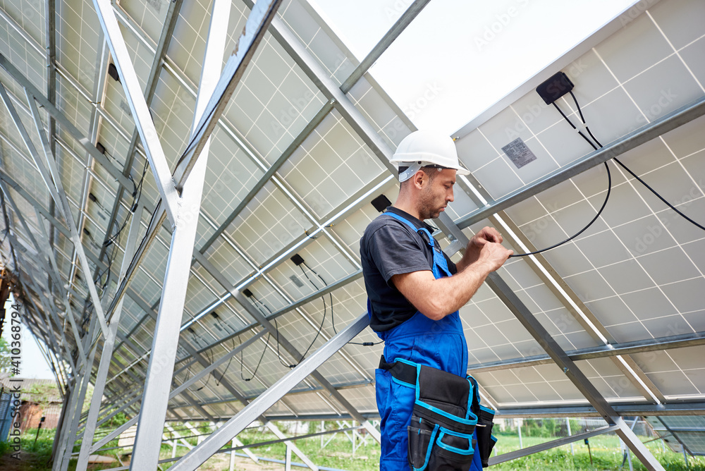 Young engineer technician making electrical wiring standing inside high exterior solar panel photo voltaic system on bright sunny summer day. Eco friendly cheap electricity generation concept.