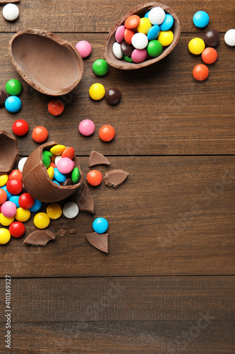 Broken chocolate eggs and colorful candies on wooden table, flat lay. Space for text