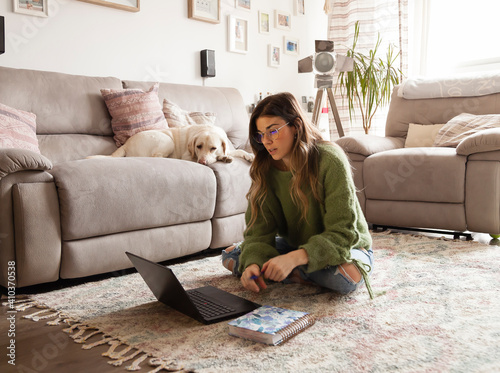Woman sitting on his living room carpet while studying or working from home with laptop, warm light and out of focus sleeping dog.
