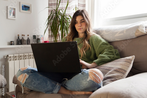 Lifestyle shot of curly blonde hair styled woman sitting confortable on a sofa while works remote from home using  his laptop with home decorated background and heating.