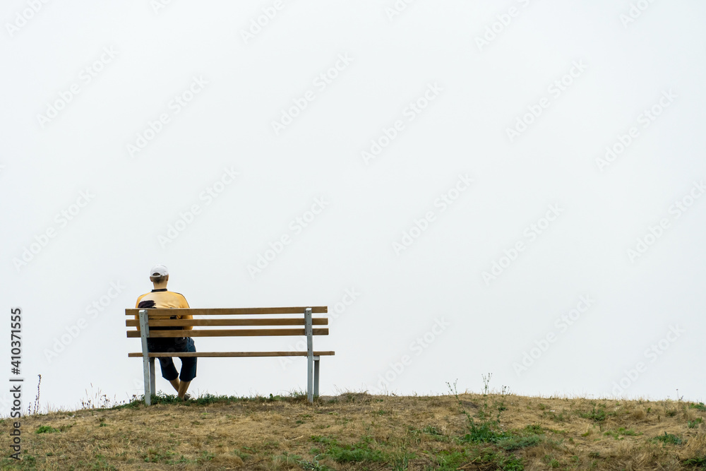 A lonely young man sits on a bench at the top of a hill against a background of thick fog. Peace and tranquility on the soul of a single man. Mourning for lost friends.