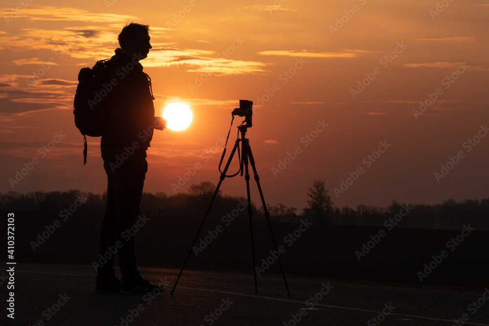 The silhouette of a person against the background of the sun and the beautiful sky. The outline of a man standing next to a tripod and a camera on the background of the sunset.