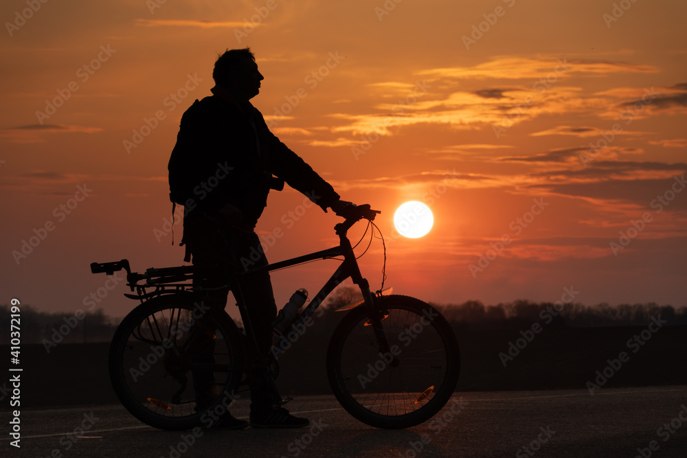 Obraz premium The silhouette of a cyclist against the background of the sun and the beautiful sky. The outline of a man standing next to his bicycle against the background of the sunset.
