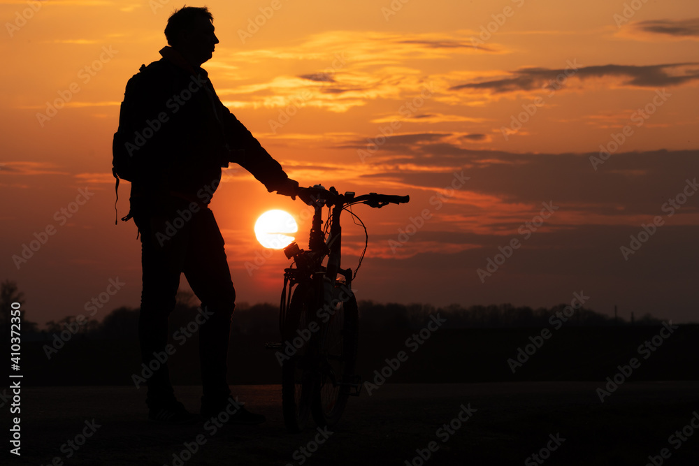 Obraz premium The silhouette of a cyclist against the background of the sun and the beautiful sky. The outline of a man standing next to his bicycle against the background of the sunset.