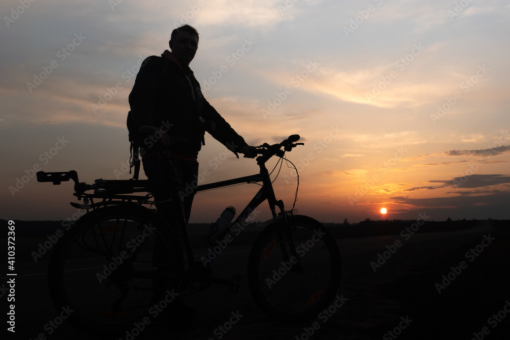 Obraz premium The silhouette of a cyclist against the background of the sun and the beautiful sky. The outline of a man standing next to his bicycle against the background of the sunset.