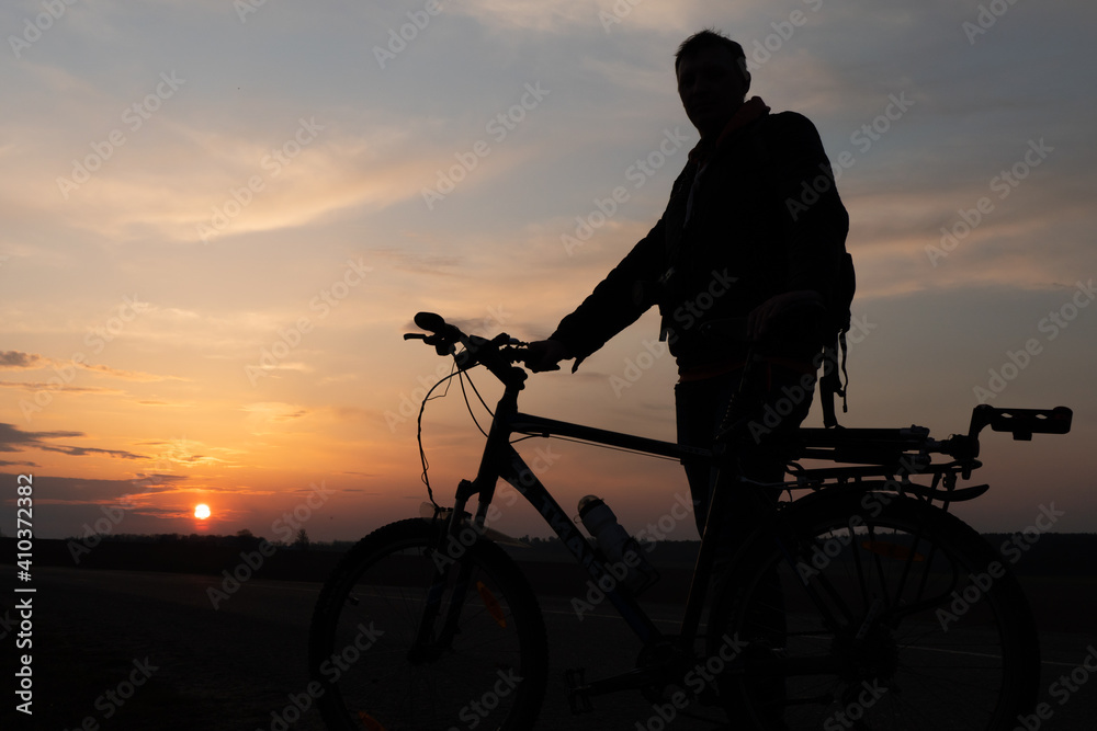 Obraz premium The silhouette of a cyclist against the background of the sun and the beautiful sky. The outline of a man standing next to his bicycle against the background of the sunset.