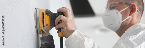 Close-up of professional worker wearing white protective face mask and working with yellow sander machine. Qualified man polish wall in apartment. Construction site and renovation concept photo