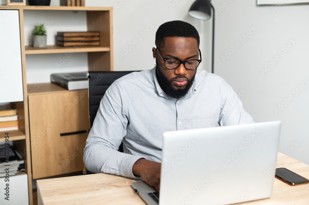 Concentrated young African-American male office worker in glasses sitting at the desk and typing on a laptop, participating in the online conference. A student stressing, taking an online exam Concentrated young African-American male office worker in glasses sitting at the desk and typing on a laptop, participating in the online conference. A student stressing, taking an online exam