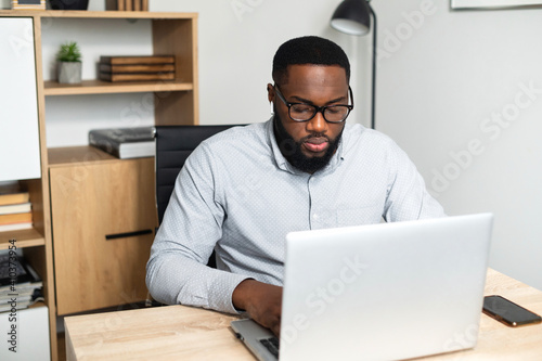 Concentrated young African-American male office worker in glasses sitting at the desk and typing on a laptop, participating in the online conference. A student stressing, taking an online exam