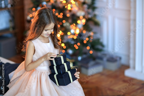 cute little girl in a pink dress holding a big box with gifts sitting near Christmas tree decorated gerlyandoy