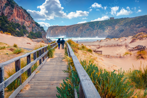 Torists walking to Cala Domestica beach on wooden pier. Windy summer scene of Sardinia, Italy, Europe. Footbridge in Canyon di Cala Domestica. Beautiful marine scenery..