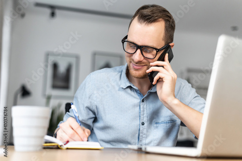A young male freelancer talking on the phone and using a laptop working remotely at home office. A guy in smart casual shirt and glasses taking notes speaking on the phone