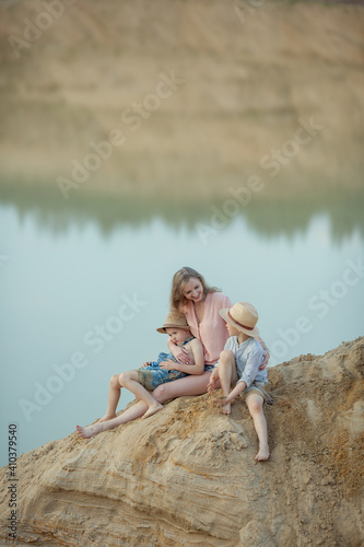 beautiful mother sits with her sons in jeans and a hat on a huge mountain at the lake