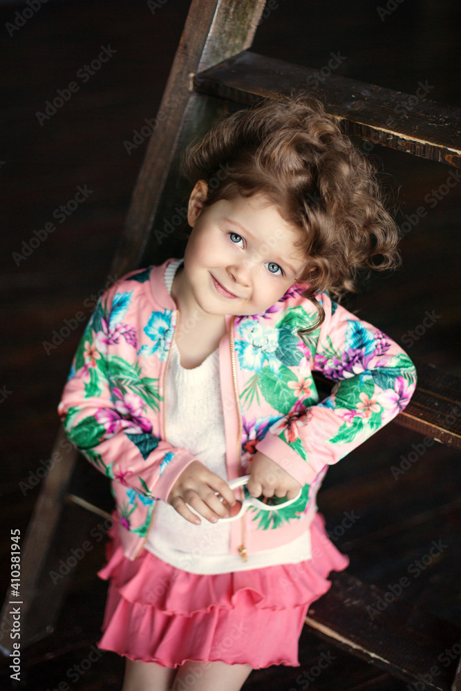 a girl with curly hair pink skirt and shirt sits on a wooden ladder.Beautiful children models. Fashion a girl with curly hair pink skirt and shirt sits on a wooden ladder.Beautiful children models. Fashion