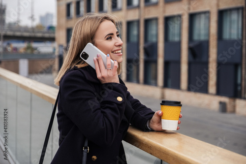 Blonde woman in dark coat drinks coffee in to go cup and works on her phone. Dark cold autumn evening. Europeaan city lights on the background. Space for text photo