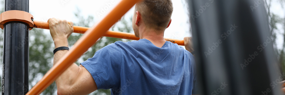 Close-up of strong male pulls up on street horizontal bar. Person in blue shirt training outdoors. Sporty guy exercise for fit body. Health and hobby. Sport and active lifestyle concept - obrazy, fototapety, plakaty 