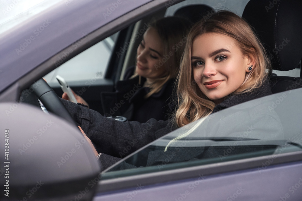 Two blonde girls in a car. One is driver and another is passenger. Dark cold autumn evening, girls wearing coats and drink coffee in to go cup