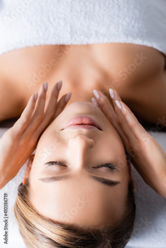 young woman with closed eyes touching face and lying on massage table in spa salon