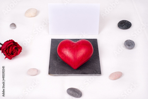 Top view on a red love heart presented as a jewel on a square slate, with a white and blank gift card behind, and stones arranged around and a red rose. Love and valentine's day 