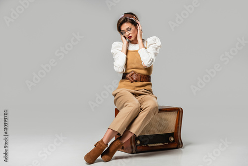 young woman in trendy retro clothes adjusting headband while sitting on vintage radio receiver on grey