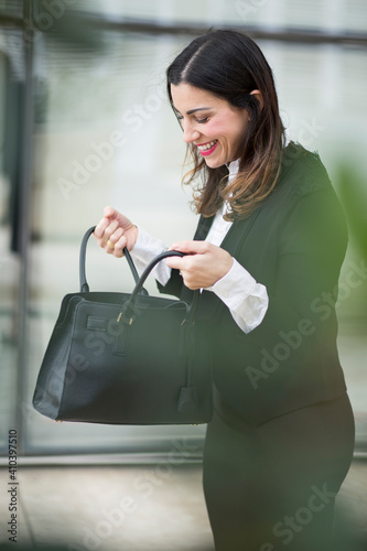 Manager donna mora, vestita in tailleur nero guarda sorridente dentro la sua borsa nera in contesto urbano moderno photo