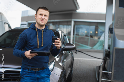 Car refueling, business, people concept. Portrait of handsome young Caucasian man in casual outfit, posing to camera with tablet and coffee to go, while his car is refueling at gas station