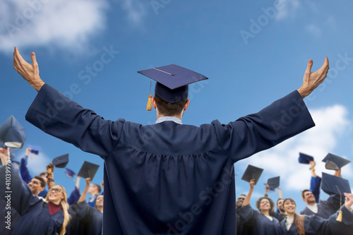 education, graduation and people concept - male graduate student over group of happy international students in bachelor gowns waving mortar boards or hats over blue sky and clouds background photo