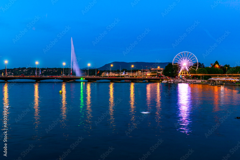 The Swiss Ferris Wheel and Pont du Mont-Blanc reflecting on Rhone river overlooking Lake Leman, French Swiss. Night view of Jet d'eau fountain and skyline of cityscape Geneva in Switzerland by evening