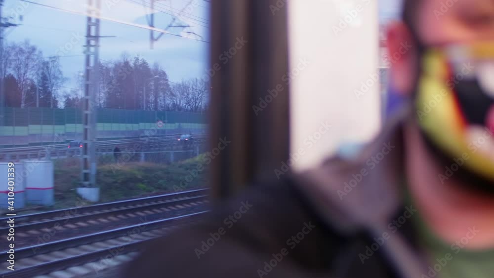 Defocused view of passanger in mask sitting ner window in wagon of train passing by road and rail way.