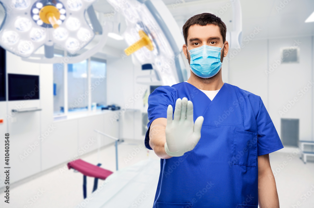 healthcare, surgery and medicine concept - doctor or male nurse in blue uniform, face medical mask for protection from virus disease and gloves showing stop gesture over hospital on background healthcare, surgery and medicine concept - doctor or male nurse in blue uniform, face medical mask for protection from virus disease and gloves showing stop gesture over hospital on background