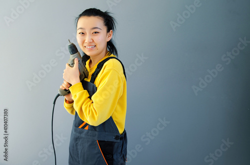 Asian Female construction worker dressed in special clothes with a cordless screwgun, a construction tool isolated on grey background