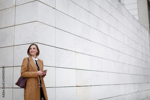 manager donna con cappotto beige , camicia bianca e cravatta nera cammina in contesto urbano utilizzando il suo smartphone sorridendo felice photo