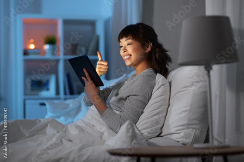 technology, internet and people concept - happy smiling young asian woman in glasses with tablet pc computer having video call in bed at home at night and showing thumbs up