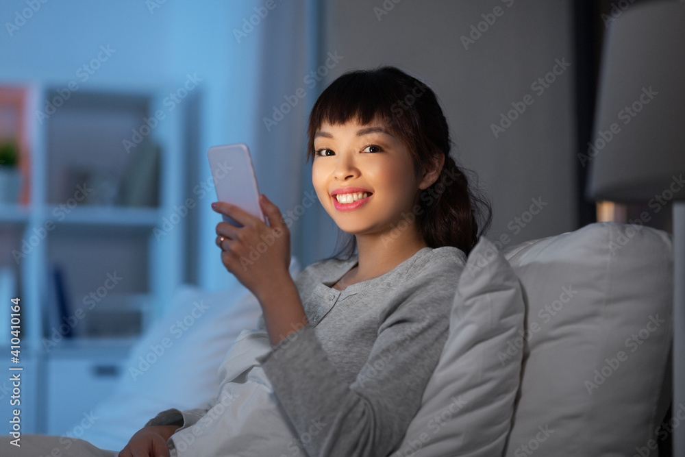 Fototapeta technology, internet, communication and people concept - happy smiling young asian woman with smartphone lying in bed at home at night