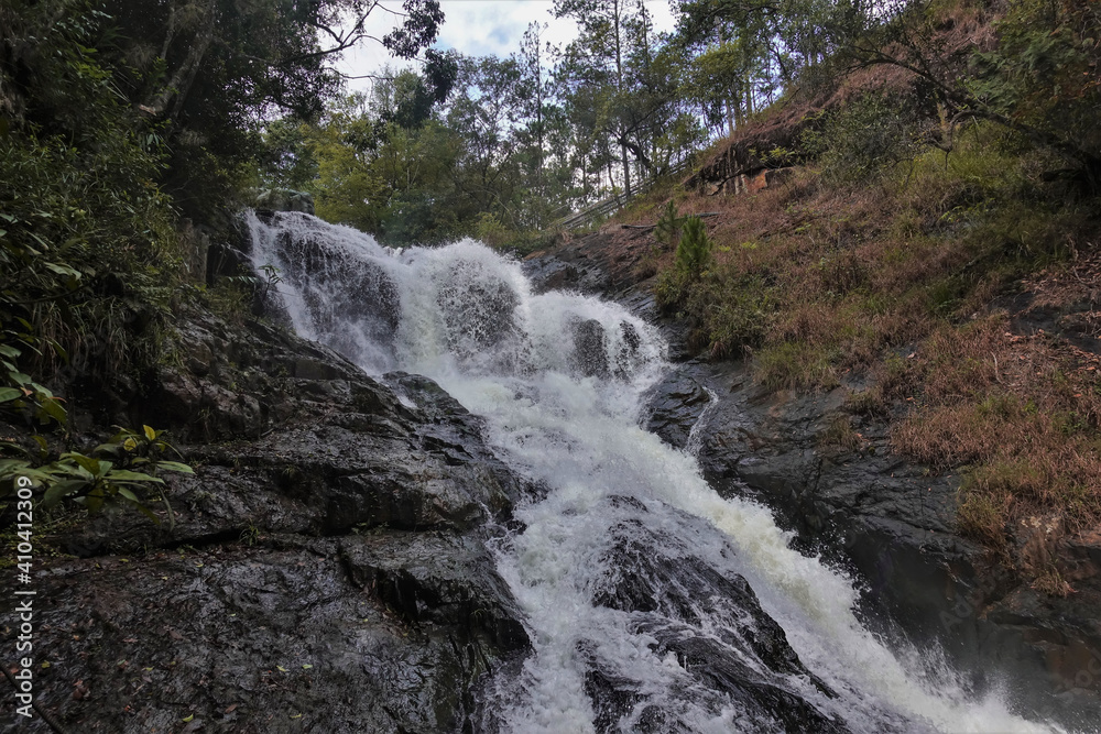 The waterfall cascades down from the rocks. Splashes and foam over water stream. Green plants all around. Vietnam. Dalat The waterfall cascades down from the rocks. Splashes and foam over water stream. Green plants all around. Vietnam. Dalat