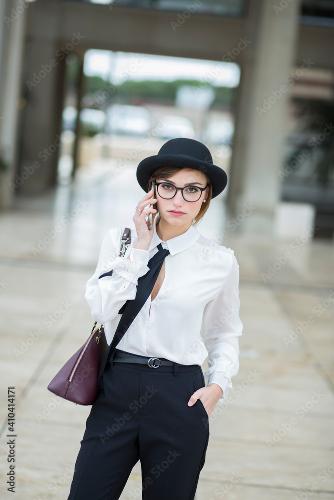 manager donna con occhiali da vista , cappello nero , camicia bianca in contesto urbano utilizzando il suo smartphone. manager donna con occhiali da vista , cappello nero , camicia bianca in contesto urbano utilizzando il suo smartphone.