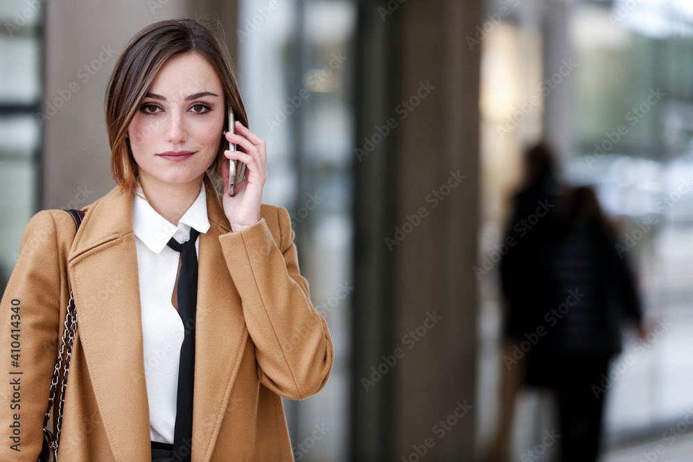 manager donna con cappotto beige , camicia bianca e cravatta nera cammina in contesto urbano utilizzando il suo smartphone - obrazy, fototapety, plakaty 