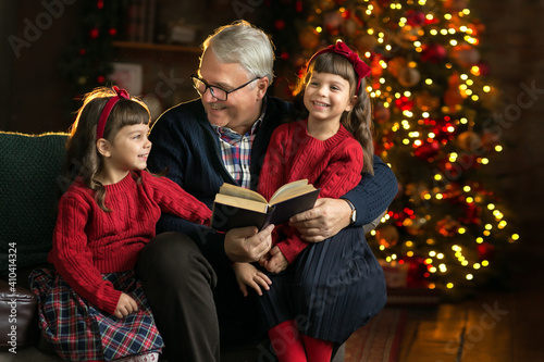 Grandfather wearing glasses, reading a book to small granddaughters twins in a room decorated for Christmas on the background of a Christmas tree. Christmas holiday concept.