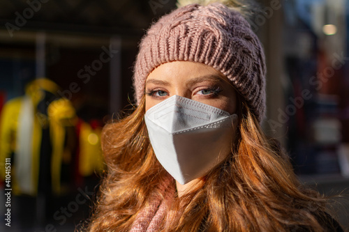 Woman with blue eyes, ginger hair, and a woollen hat, wearing a protective face mask (FFP-2) during pandemic isolation as part of the prevention of COVID-19 diseases. City street scene.