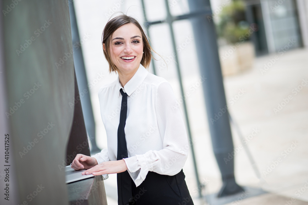 Foto Stock manager donna bionda con i capelli a caschetto, camicia bianca e cravatta  nera, lavora al computer in esterni e sorride felice | Adobe Stock