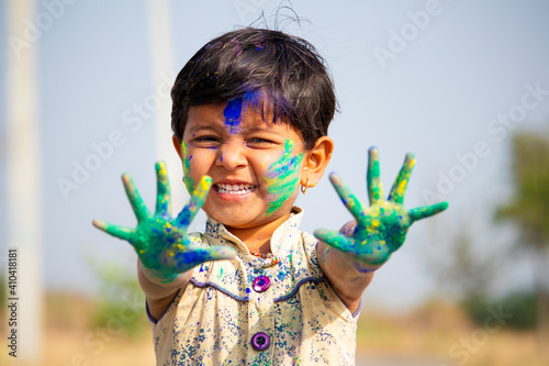 young cute cheerful little girl kid with applied holi colors powder showing colorful hands to camera during holi festival celebration.