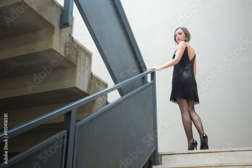 Bellissima ragazza con i capelli a caschetto e un abito nero corto con bretelle scende dalle scale con espressione seria photo