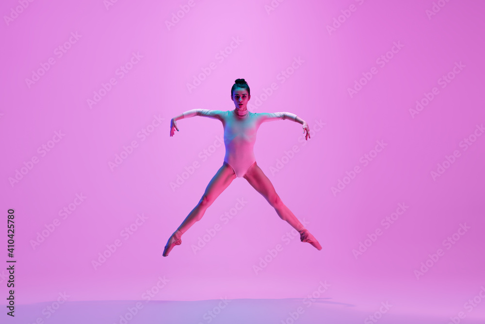 Flying on. Young and graceful ballet dancer on pink studio background in neon light. Art, motion, action, flexibility, inspiration concept. Flexible caucasian ballet dancer, moves in glow.